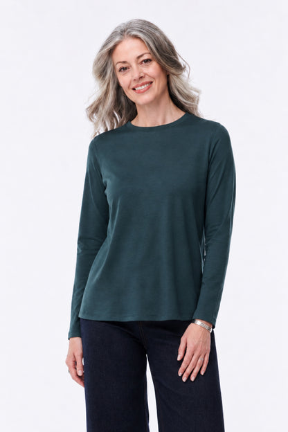 A woman with gray hair wearing a dark teal-green long-sleeve crewneck top and dark pants, standing against a plain white studio background.