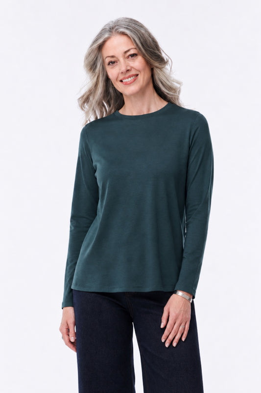 A woman with gray hair wearing a dark teal-green long-sleeve crewneck top and dark pants, standing against a plain white studio background.