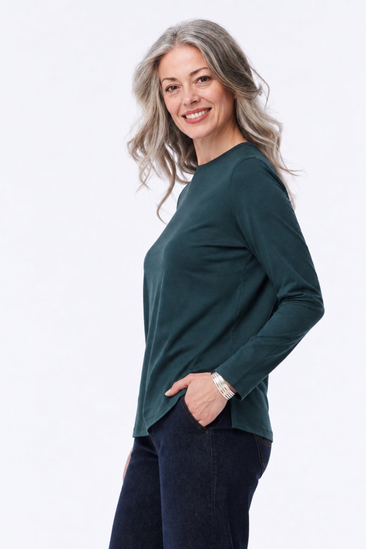 A smiling woman with gray hair is shown in profile wearing a dark green long-sleeve top and dark blue jeans against a plain white studio background.
