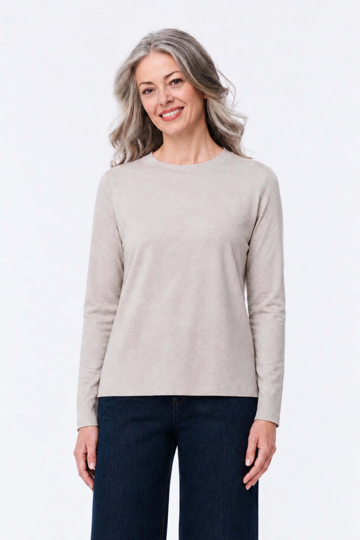 A woman with gray hair wearing a light beige long-sleeve crewneck sweater and dark blue jeans, standing against a plain white studio background.