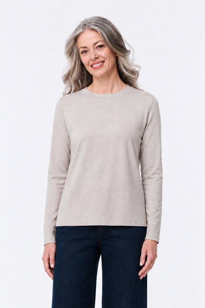 A woman with gray hair wearing a light beige long-sleeve crewneck sweater and dark blue jeans, standing against a plain white studio background.