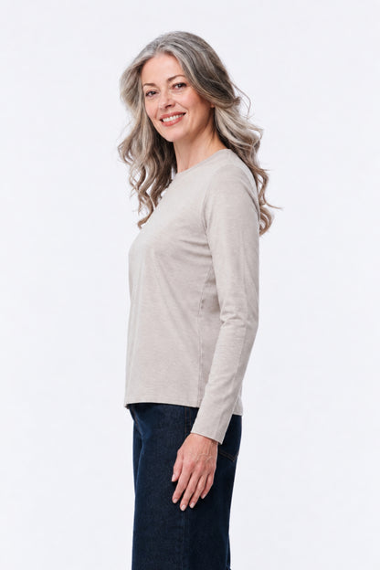 A woman with wavy gray hair is shown in profile wearing a light beige long-sleeve knit top and dark blue jeans against a plain white studio background.