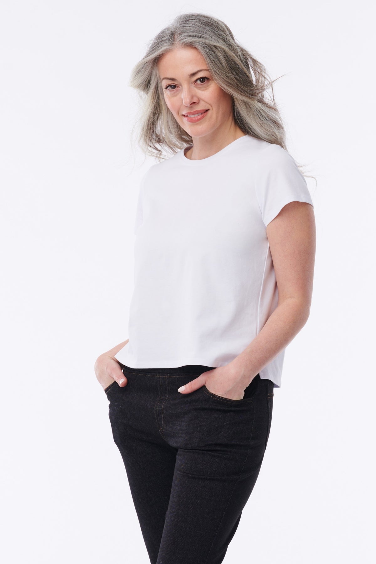 A woman with gray hair wearing a white short-sleeve crewneck t-shirt and black jeans, standing against a plain white studio background.