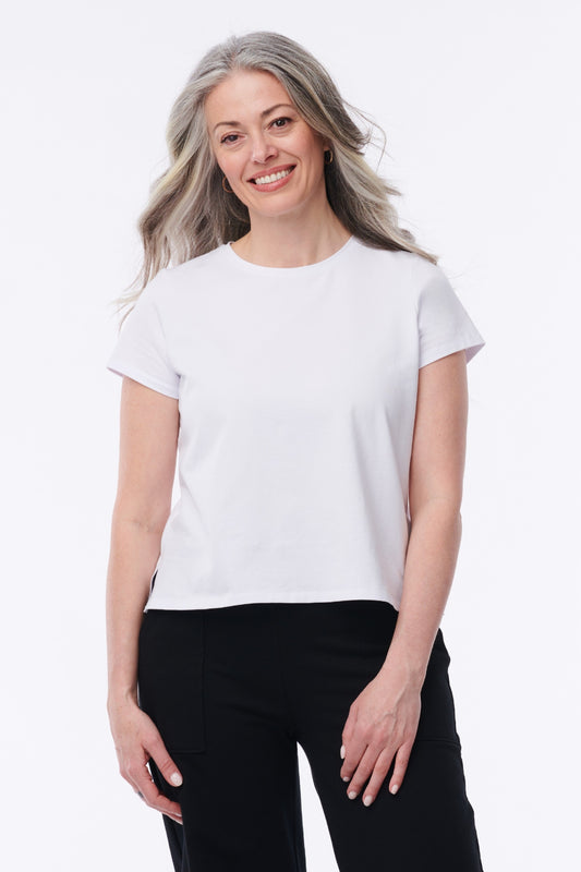 A smiling woman with gray hair wearing a plain white short-sleeve crewneck t-shirt and black pants, standing against a solid white studio background.