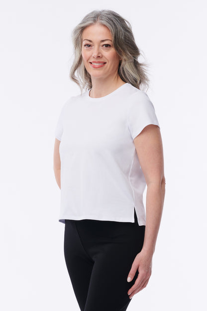 A woman with gray hair wearing a white short-sleeve crewneck top and black leggings, standing against a plain white studio background.