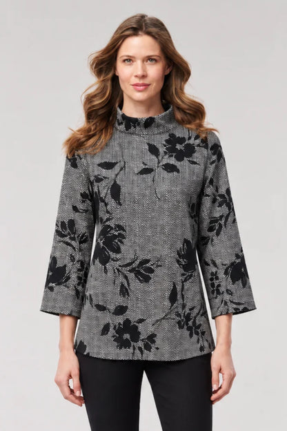 A woman modeling a gray and black floral patterned mock-neck top with three-quarter sleeves, paired with black pants, against a light gray studio background.
