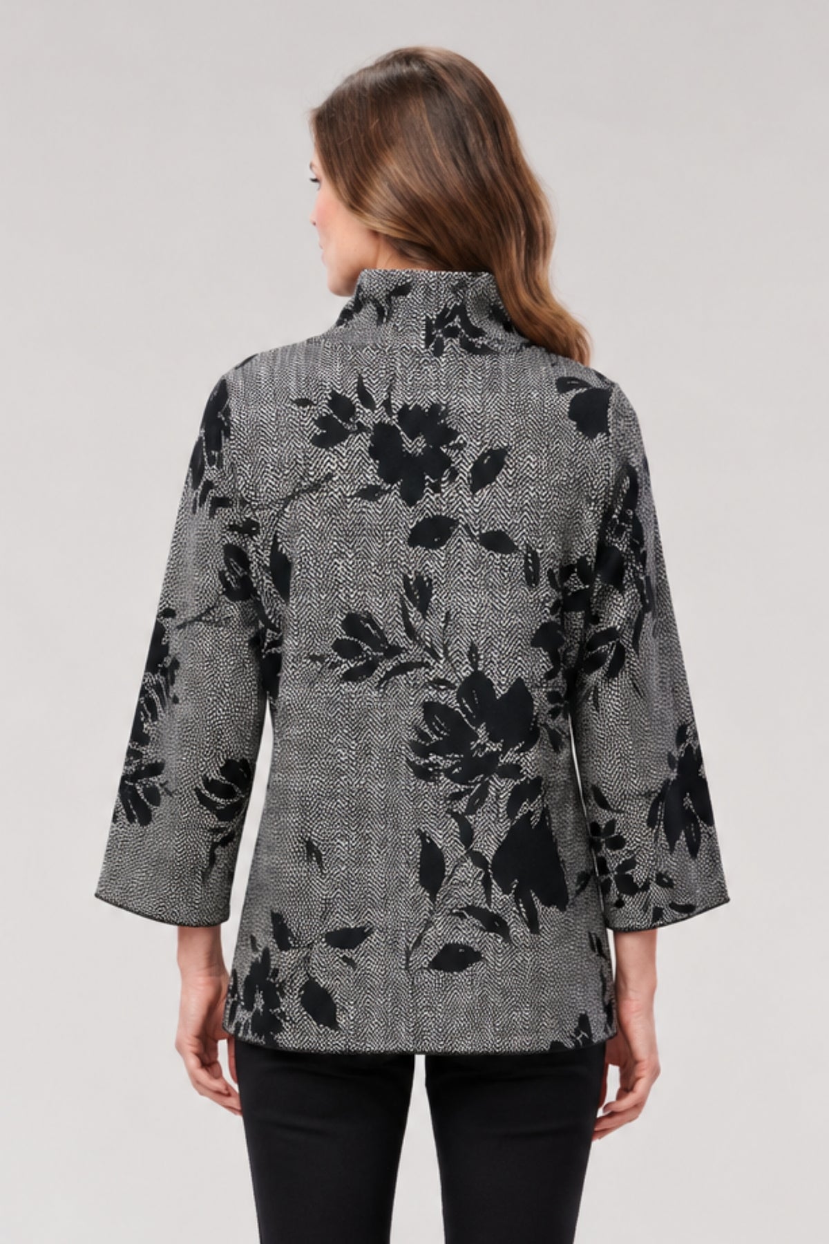 A woman viewed from the back wearing a gray textured jacket with a black floral pattern, paired with black pants, against a light gray studio background.