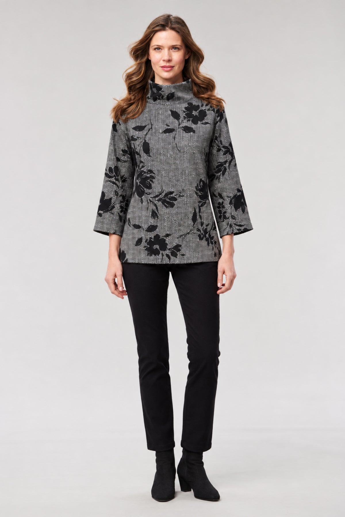 A woman modeling a gray and black floral patterned mock-neck long-sleeve top, paired with black pants and black ankle boots, against a light gray studio background.