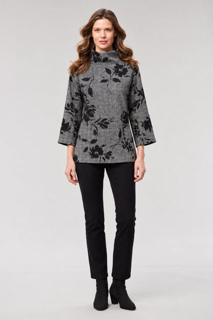 A woman modeling a gray and black floral patterned mock-neck long-sleeve top, paired with black pants and black ankle boots, against a light gray studio background.