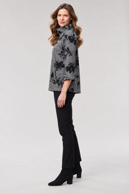 A woman modeling a gray and black floral patterned turtleneck top, paired with black pants and black heeled ankle boots, against a light gray studio background.