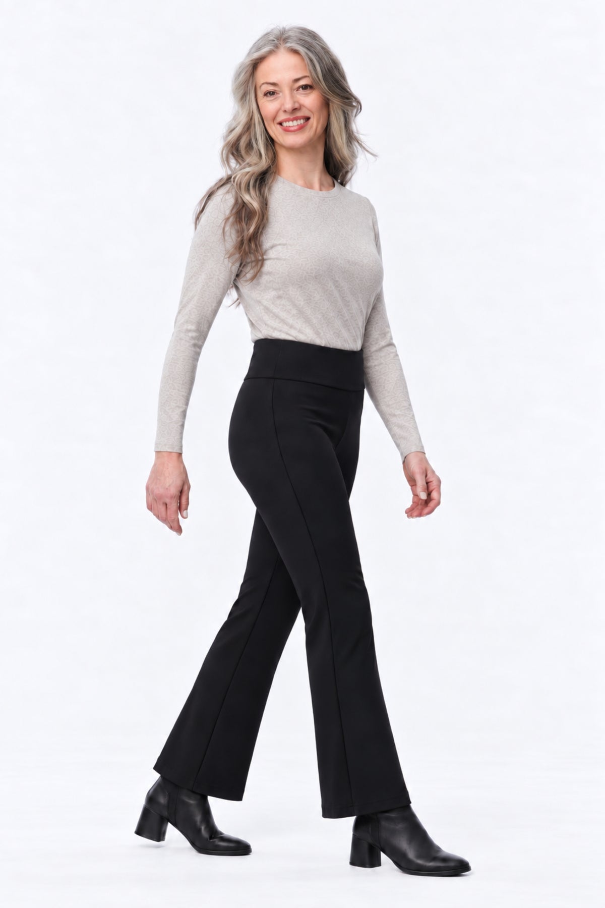 A woman modeling black flared pants with a light beige long-sleeve knit top and black ankle boots, standing against a plain white studio background.