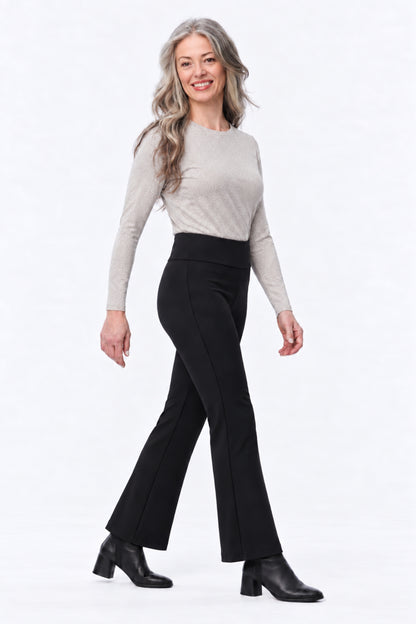 A woman modeling black flared pants with a light beige long-sleeve knit top and black ankle boots, standing against a plain white studio background.