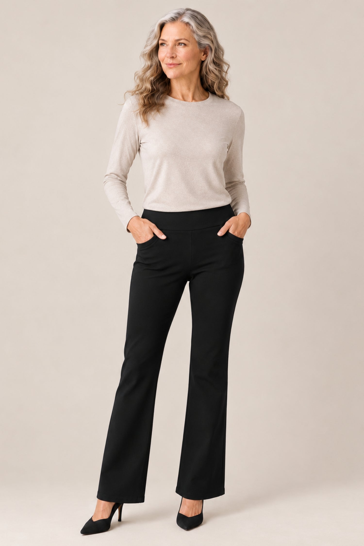 A woman modeling black flared pants with a light beige long-sleeve top, standing against a plain beige studio background.