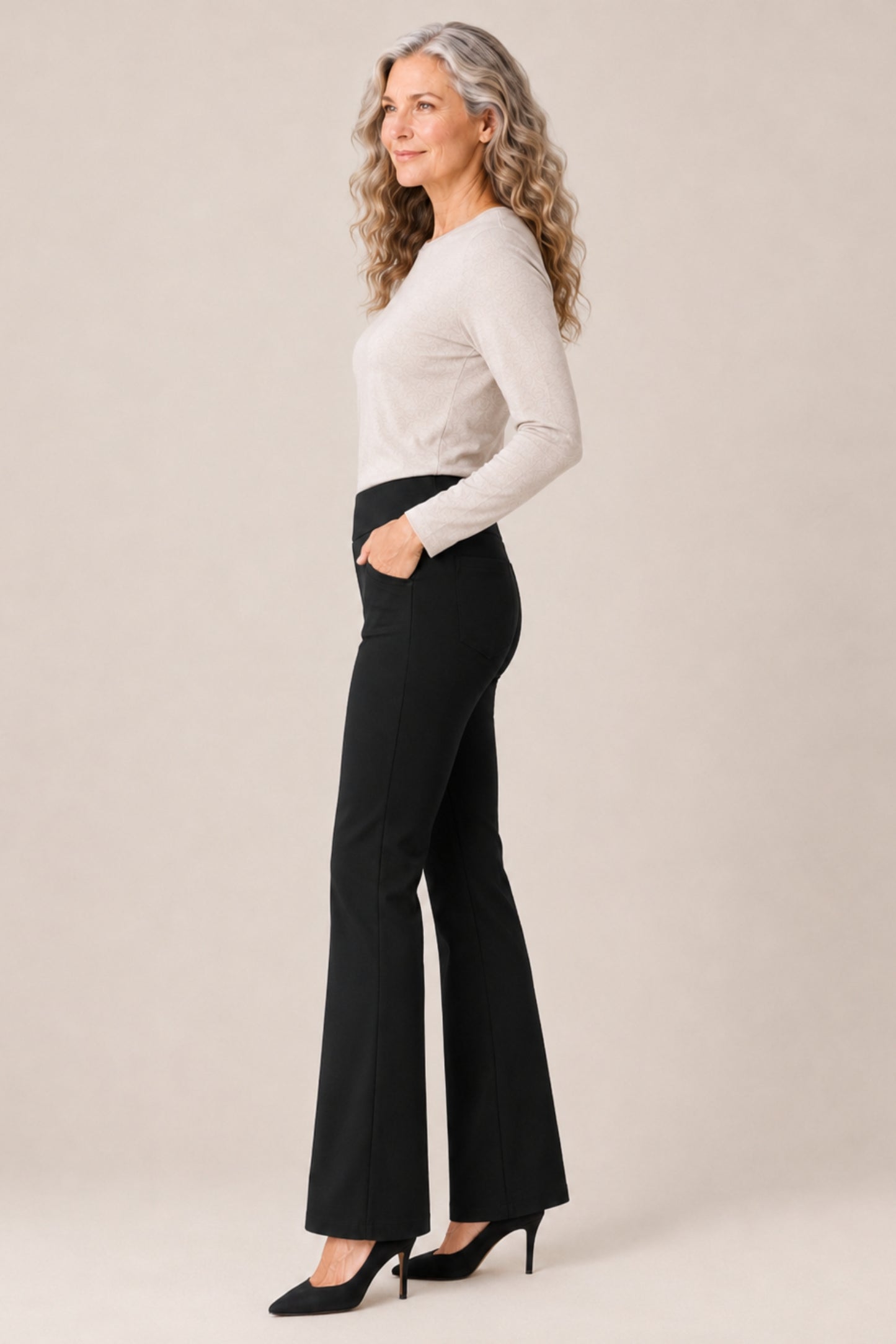 A woman with long wavy gray hair stands in profile wearing a fitted light beige long-sleeve top, black flared pants, and black heels against a plain warm beige studio background.