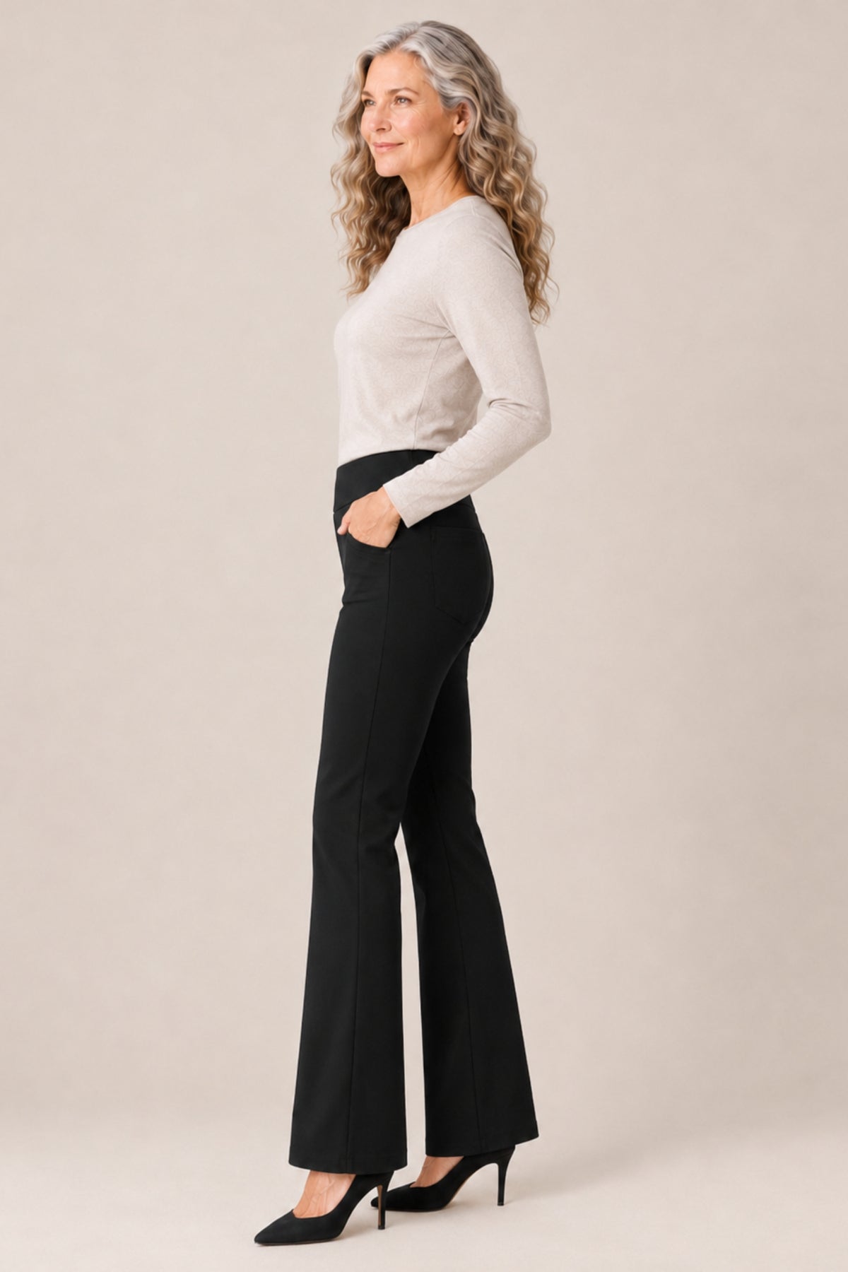 A woman with long wavy gray hair stands in profile wearing a fitted light beige long-sleeve top, black flared pants, and black heels against a plain warm beige studio background.