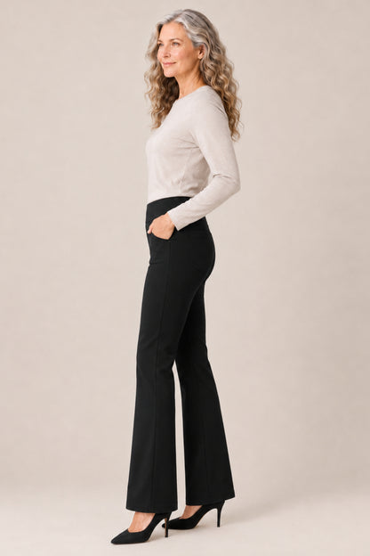 A woman with long wavy gray hair stands in profile wearing a fitted light beige long-sleeve top, black flared pants, and black heels against a plain warm beige studio background.