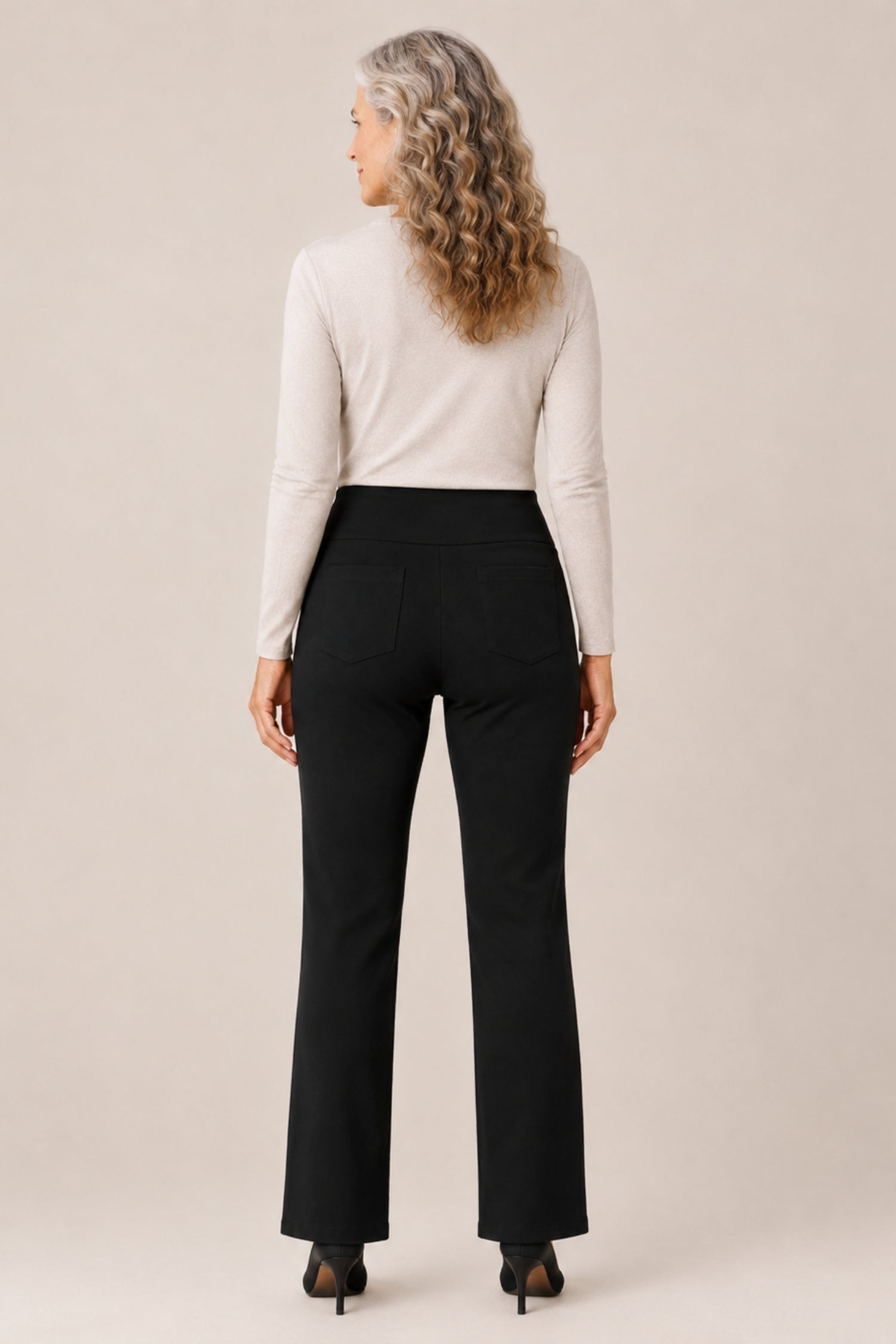 A woman with long, wavy blonde hair is shown from the back wearing a cream mock-neck long-sleeve top and black high-waisted flared pants, paired with black heels, against a solid light beige studio background.