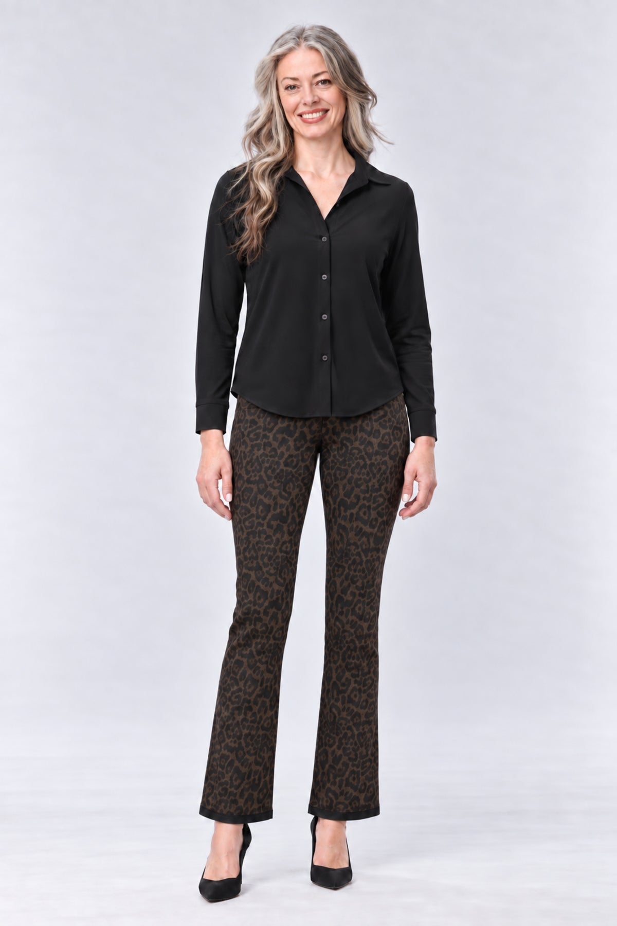 A woman modeling a black long-sleeve button-up shirt and brown leopard-print flared pants against a light gray studio background.