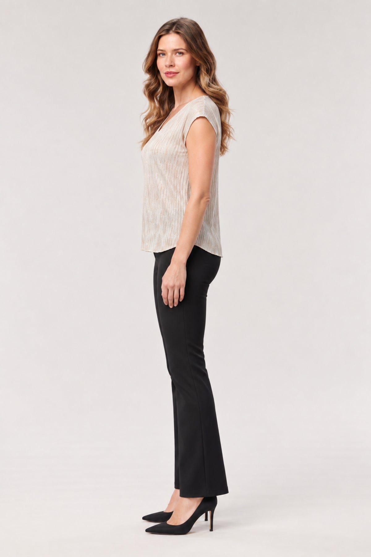 A woman modeling a sleeveless textured top in a light beige/cream color paired with black flared pants and black heels, standing against a plain light gray studio background.