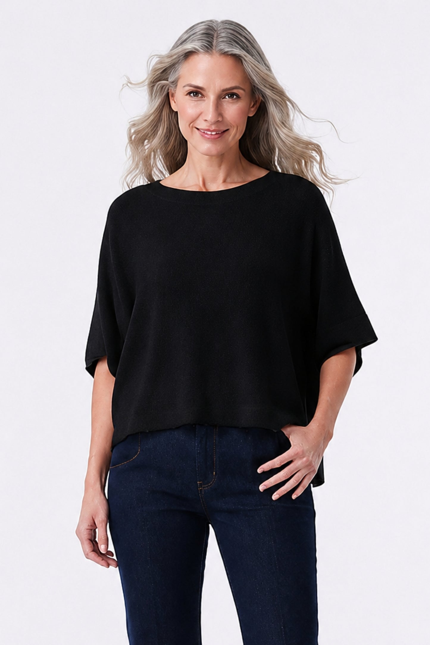 A woman with blonde hair wearing a black short-sleeve boat-neck top and dark blue jeans, standing against a light gray studio background.