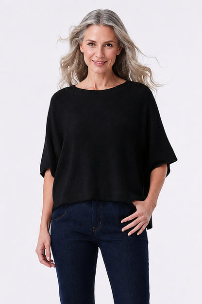 A woman with blonde hair wearing a black short-sleeve boat-neck top and dark blue jeans, standing against a light gray studio background.