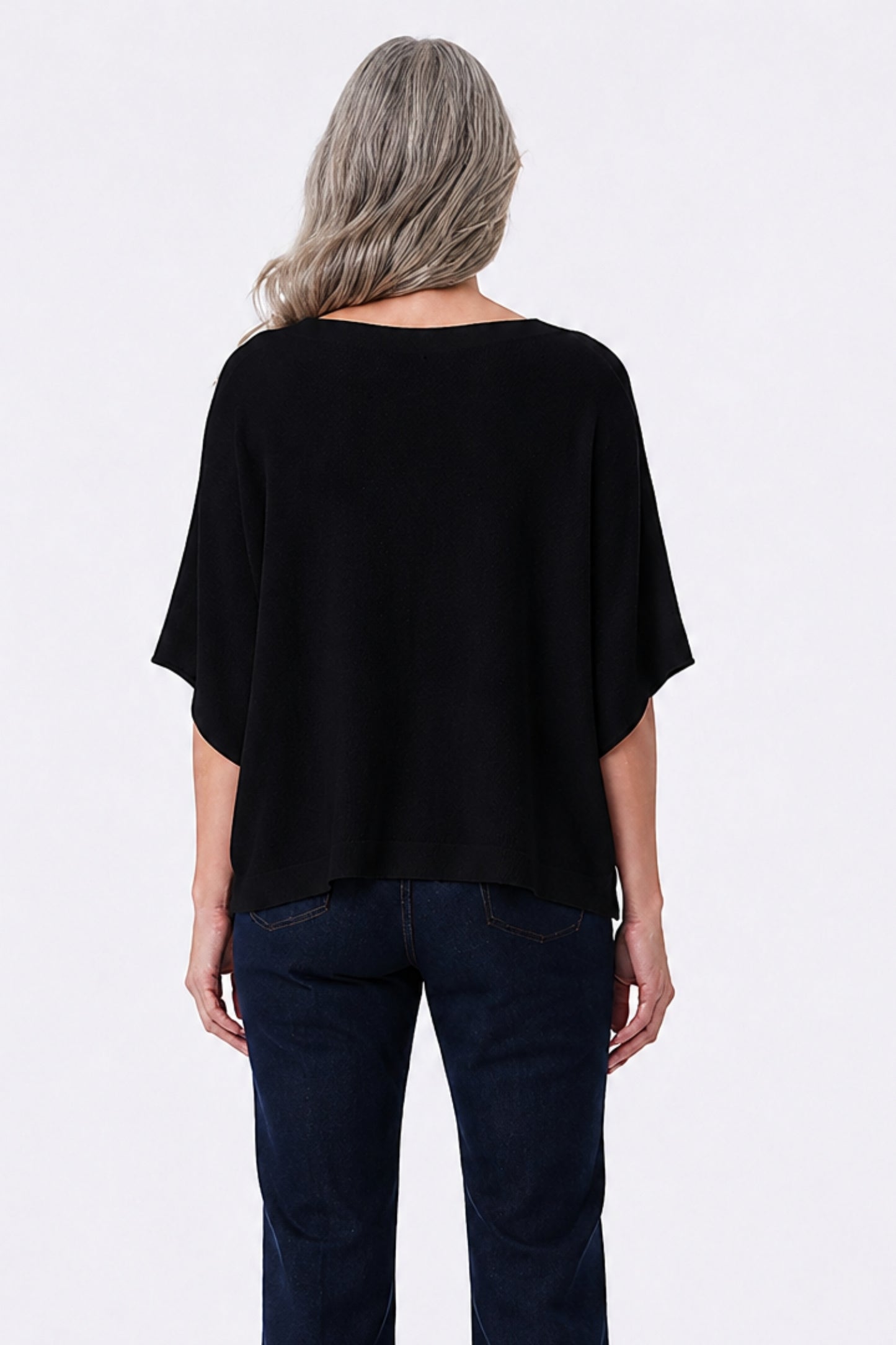 A woman viewed from the back wearing a loose black short-sleeve top and dark blue jeans against a plain light gray studio background.