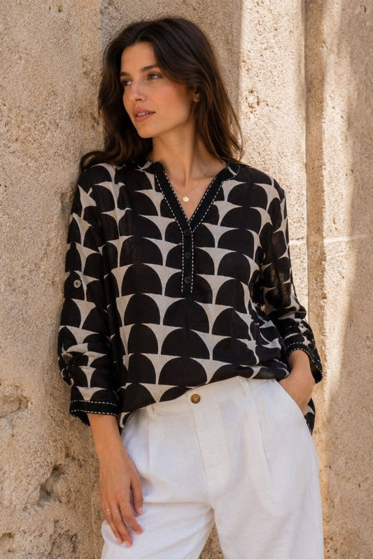 A woman wearing a black and beige geometric pattern blouse with white pants, leaning against a textured stone wall.