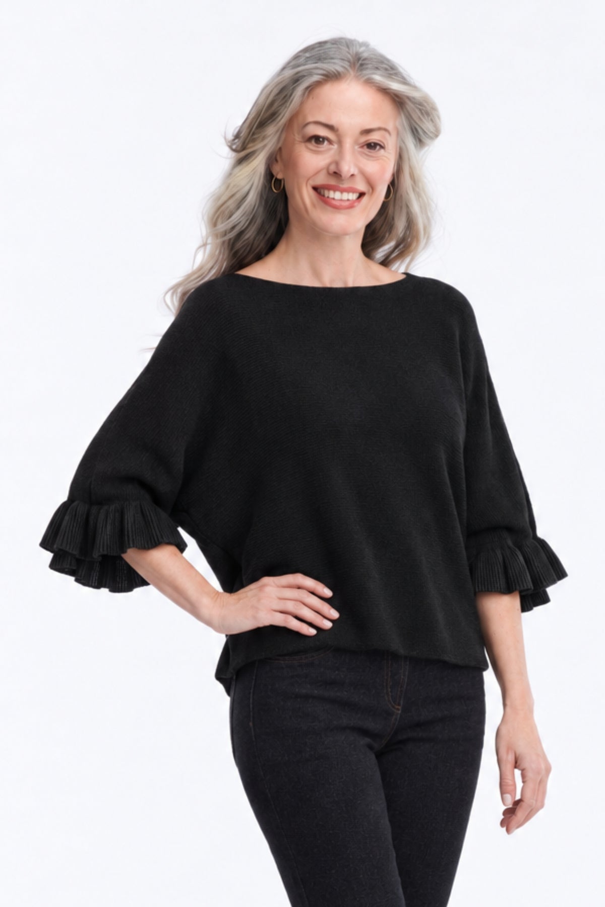 A smiling woman with gray hair wearing a black boat-neck knit top with ruffled elbow-length sleeves and black pants, standing against a plain white studio background.