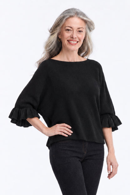 A smiling woman with gray hair wearing a black boat-neck knit top with ruffled elbow-length sleeves and black pants, standing against a plain white studio background.