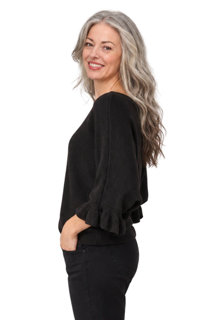A woman with long gray hair is shown from the side/back wearing a black knit top with ruffled three-quarter sleeves and black pants, photographed against a plain white studio background.