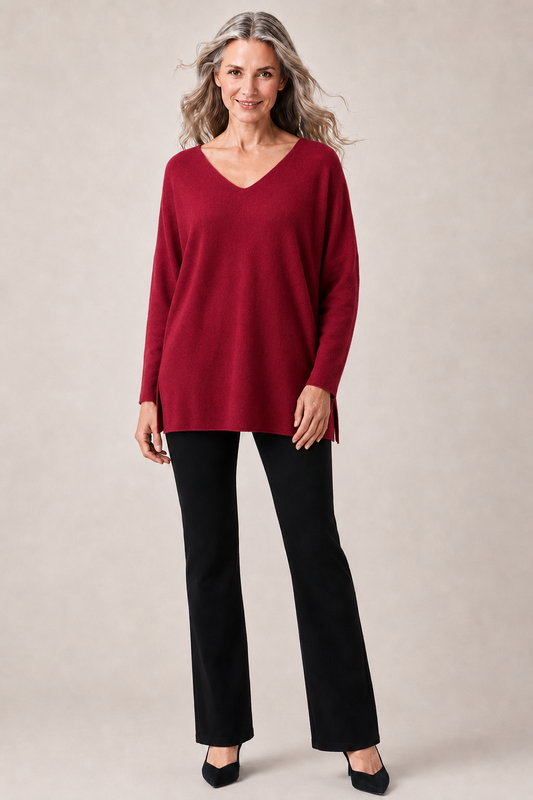 A woman modeling a long-sleeve V-neck knit top in deep burgundy, paired with black pants and black heels, against a plain beige studio background.