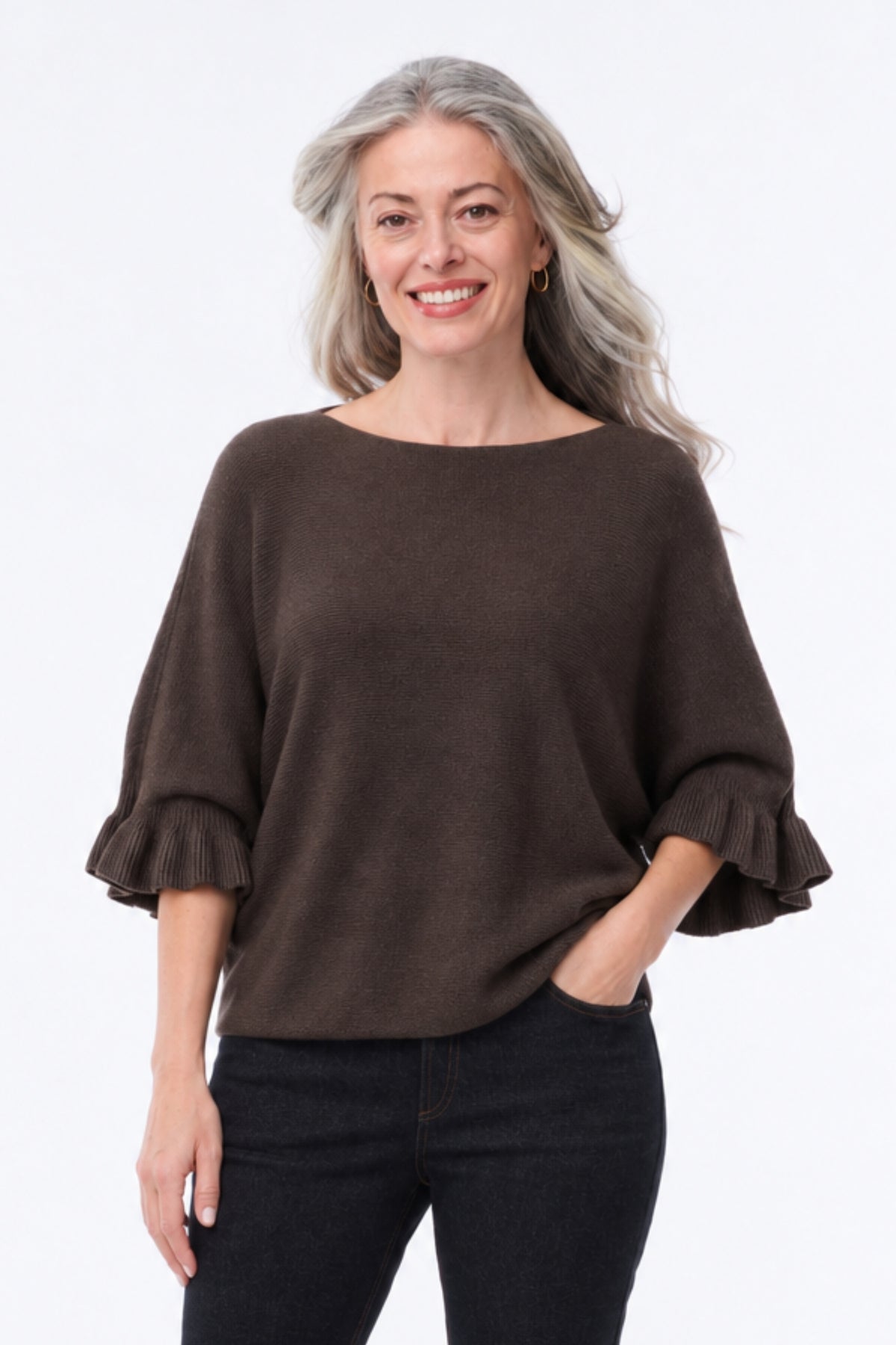 A smiling woman with gray hair wearing a dark brown boat-neck knit top with ruffled elbow-length sleeves and black pants, standing against a plain white studio background.