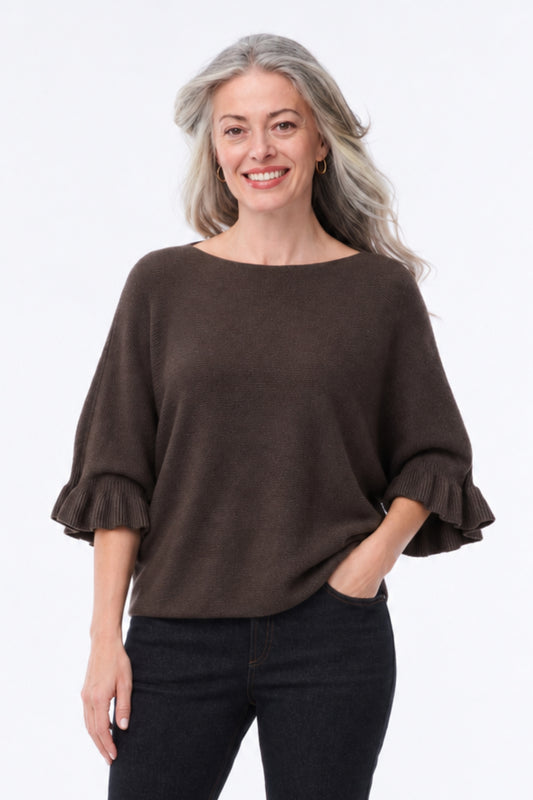 A smiling woman with gray hair wearing a dark brown boat-neck knit top with ruffled elbow-length sleeves and black pants, standing against a plain white studio background.
