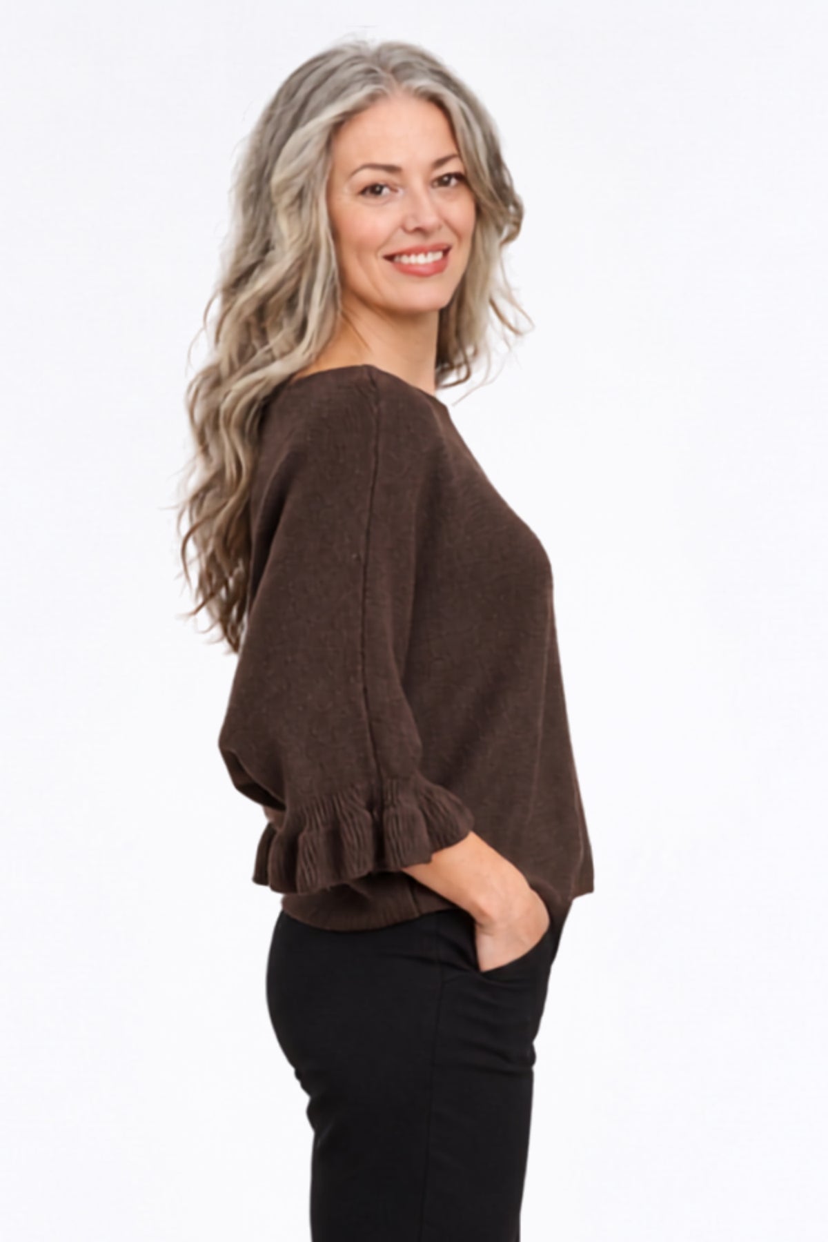 A woman with long wavy gray hair is shown from the side/back wearing a brown knit sweater with ruffled cuffs and black pants, photographed against a plain white studio background.