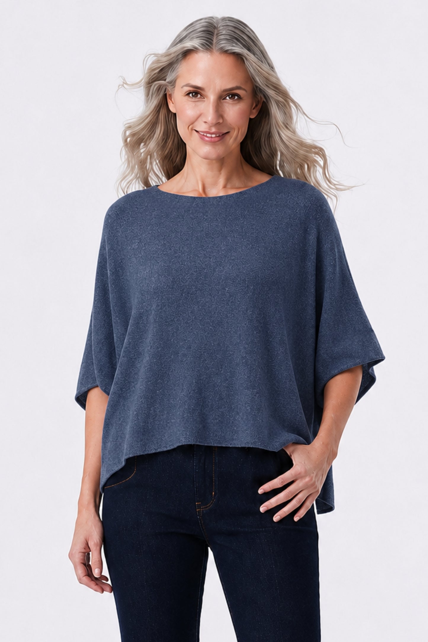 A woman modeling a slate blue knit top with a wide boat neckline and short wide sleeves, paired with dark jeans, against a light gray studio background.