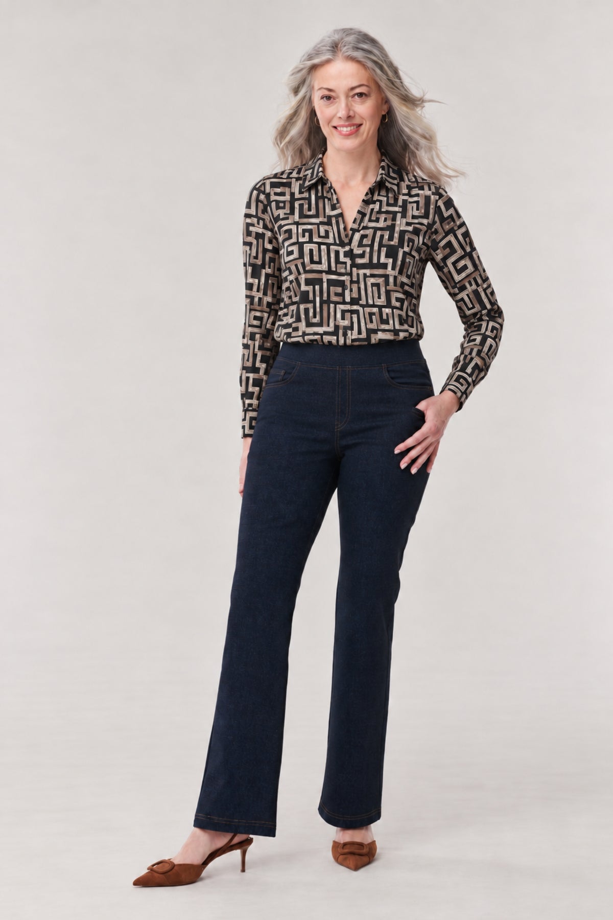 A woman modeling a long-sleeve button-up blouse with a black and beige geometric maze pattern, paired with dark blue flared jeans and brown pointed-toe heeled mules, against a light gray studio background.