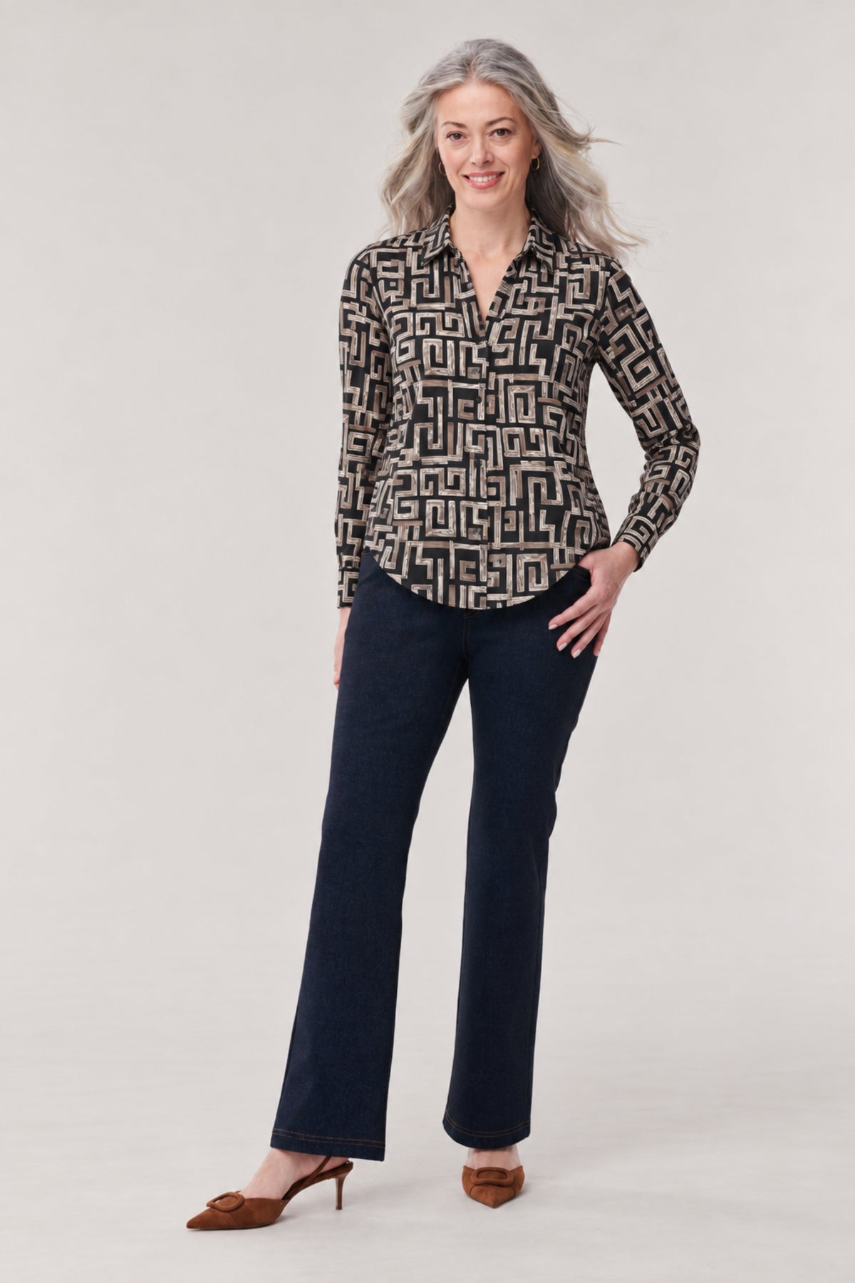 A woman modeling a long-sleeve button-up blouse with a black and beige geometric maze pattern, paired with dark blue jeans and brown pointed-toe heels, against a light gray studio background.
