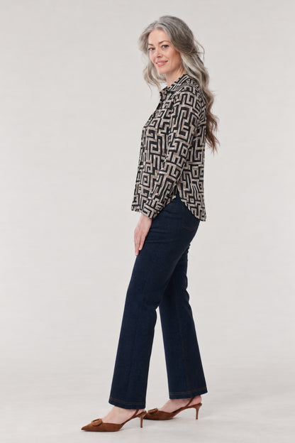 A woman modeling a black and beige geometric patterned long-sleeve blouse with dark blue flared jeans and brown slingback heels, standing against a light gray studio background.