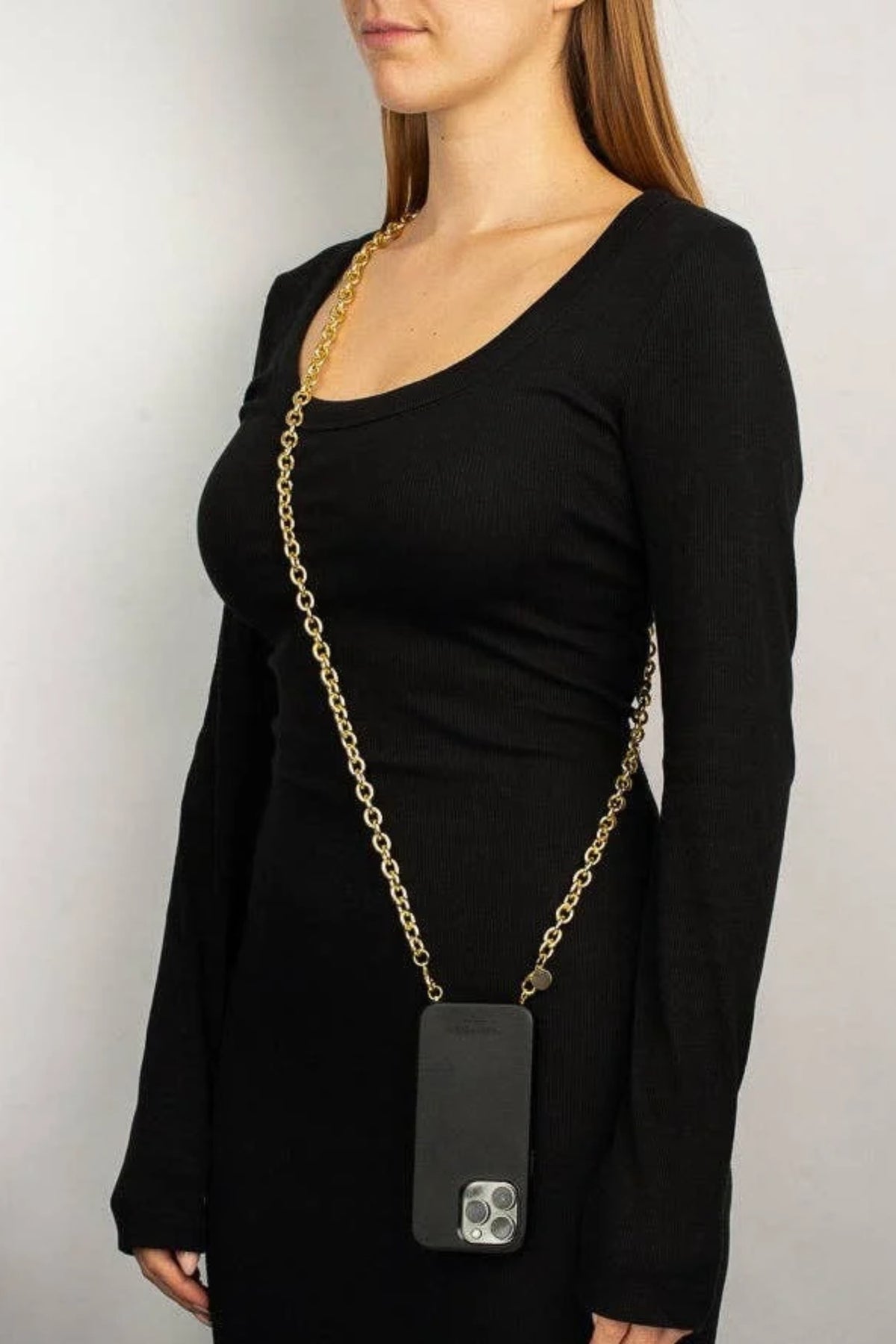 A woman wearing a black long-sleeve top and black dress, wearing a gold chain crossbody bag with a black pouch over her shoulder, against a light gray background.