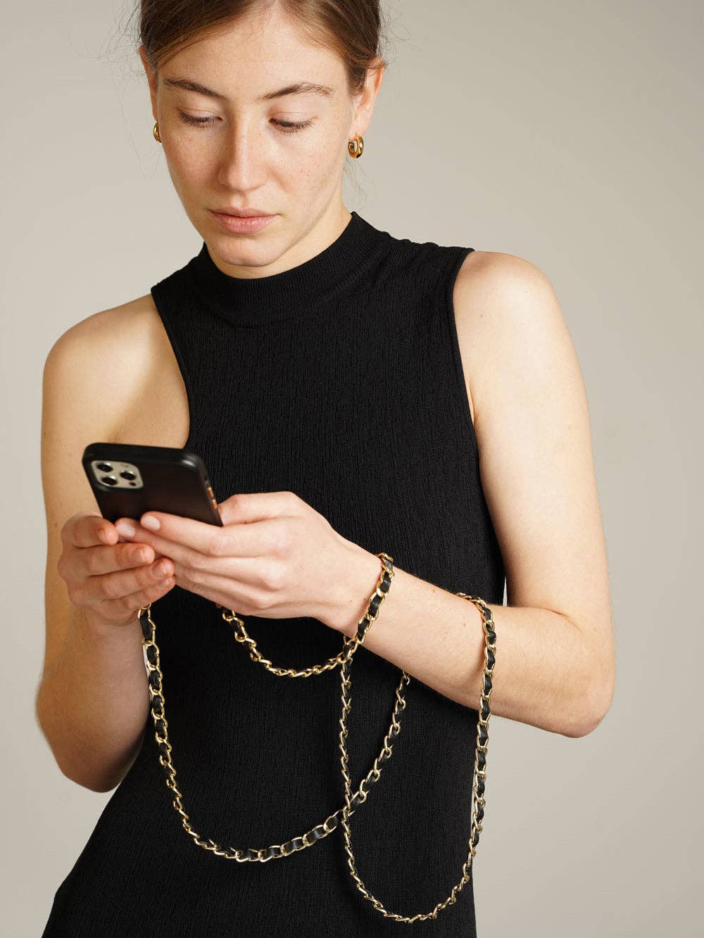A woman in a black sleeveless mock-neck top looks down at a black smartphone she is holding. A gold-tone chain accessory with black links drapes over her arms and torso.
