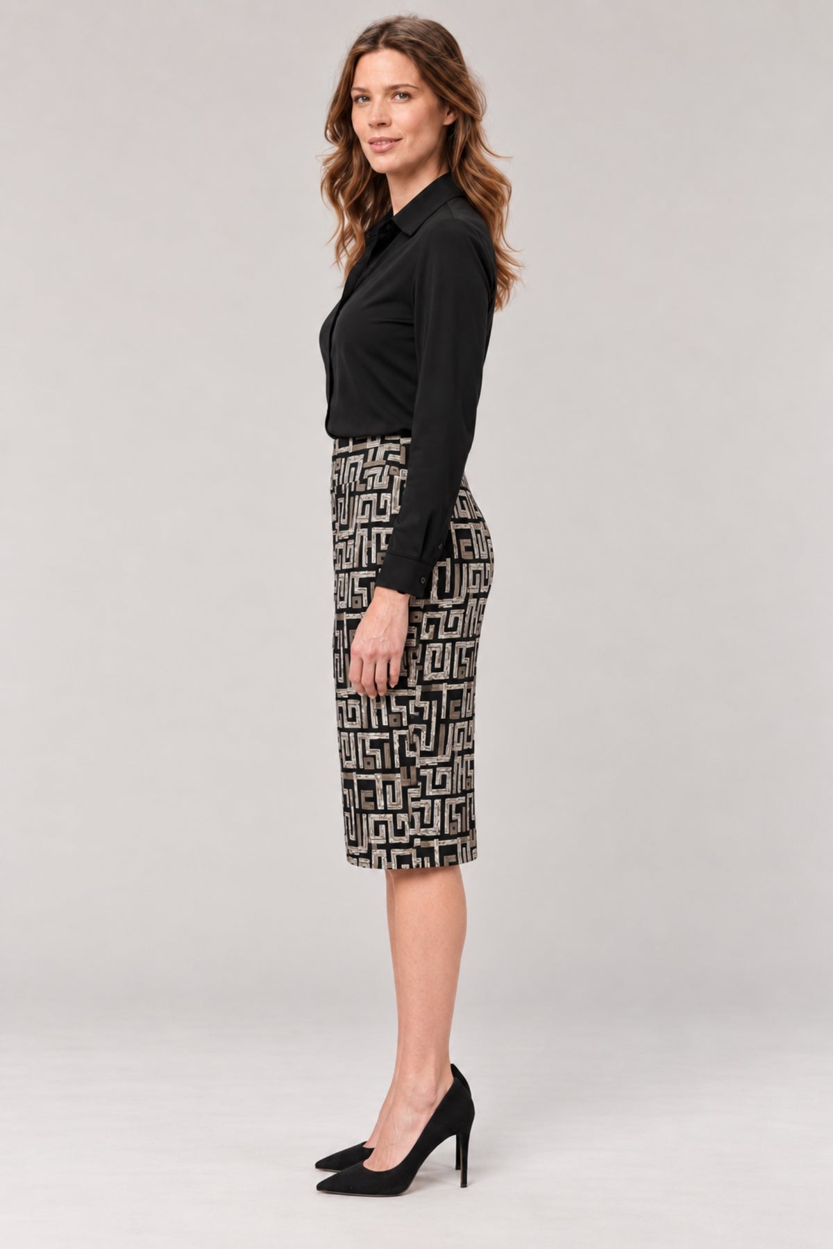 A woman stands in profile wearing a black long-sleeve blouse and a black-and-white geometric patterned knee-length skirt with black high heels, against a light gray studio background.