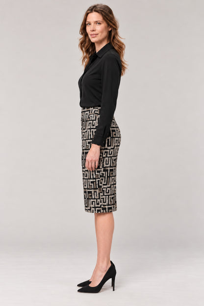 A woman stands in profile wearing a black long-sleeve blouse and a black-and-white geometric patterned knee-length skirt with black high heels, against a light gray studio background.
