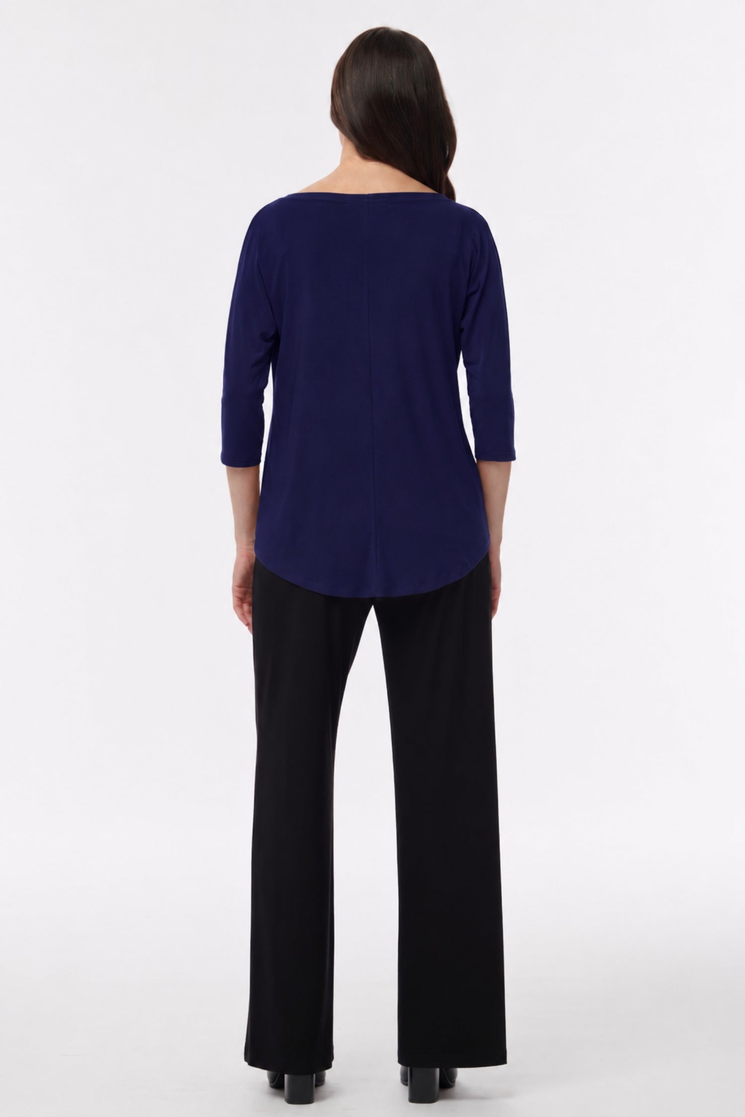 A woman viewed from the back wearing a navy blue three-quarter sleeve top and black wide-leg trousers, standing against a plain light gray studio background.